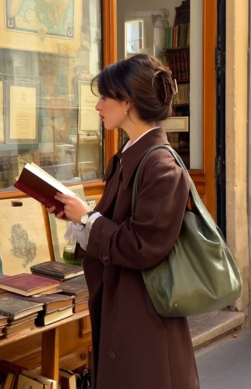 Woman in a brown coat holding a green bag and reading a book outside a bookstore.