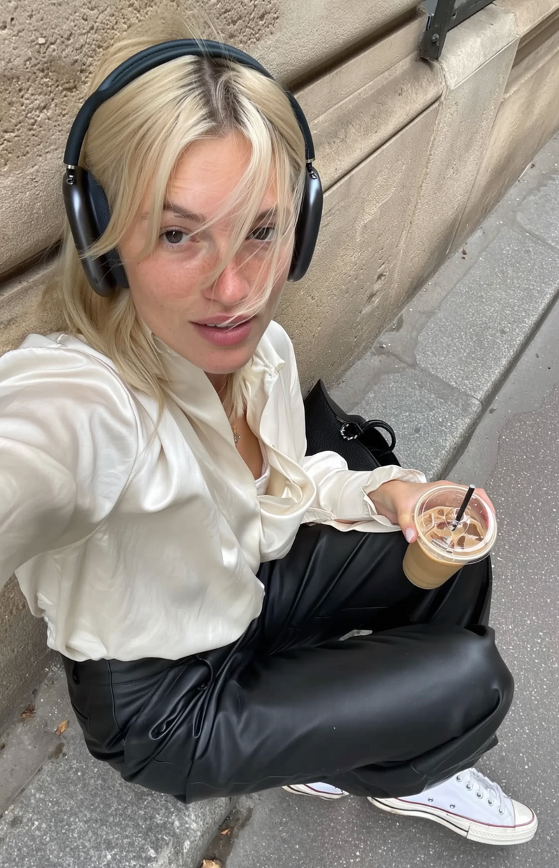Person sitting on a step with headphones and a drink, wearing a white shirt and black pants.